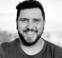 Homem branco, soridente, de meia idade, de aproximadamente 36 anos, 
 cabelos curtos, crespo e escuros, olhos castanhos de barbas escuras de tamanho média, 
 vestindo um t-shert azul.